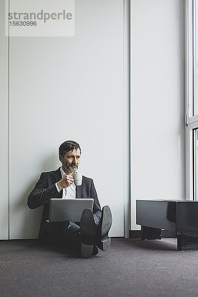 Reifer Geschäftsmann im Büro  der mit Kaffeetasse und Laptop auf dem Boden sitzt