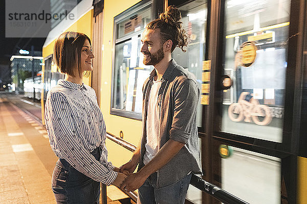 Lächelndes Paar  das sich an den Händen hält und sich mit der Straßenbahn im Hintergrund anschaut  Berlin  Deutschland