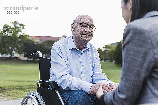 Porträt eines älteren Mannes  der im Rollstuhl sitzt und mit seiner Enkelin Händchen hält