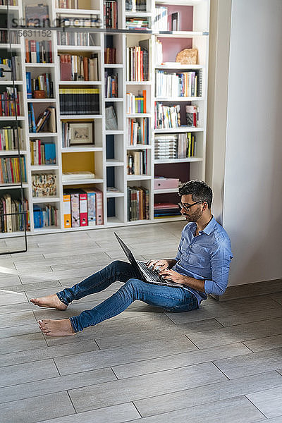 Barfüssiger junger Mann sitzt zu Hause mit Laptop auf dem Boden