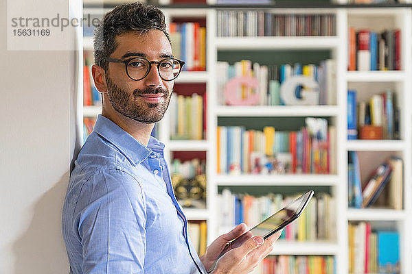 Porträt eines lächelnden jungen Mannes  der zu Hause mit einem digitalen Tablet vor einem Bücherregal steht