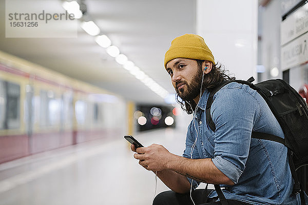 Mann mit Rucksack hört mit Smartphone und Kopfhörern Musik  während er am Bahnsteig wartet  Berlin  Deutschland
