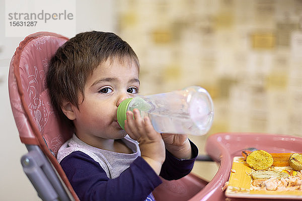 Porträt eines kleinen Jungen  der auf einem Hochstuhl sitzt und Wasser aus einer Flasche trinkt