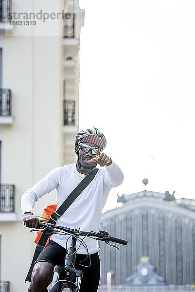 Stilvoller junger Mann mit Fahrrad und Kuriertasche in der Stadt  der mit dem Finger zeigt