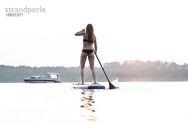 Rückenansicht einer jungen Frau  die bei Sonnenuntergang beim Paddelboarden aufsteht  Starnberger See  Deutschland