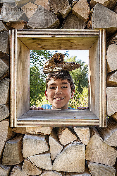 Porträt eines lächelnden Jungen  der durch ein Fenster im Holzstapel schaut