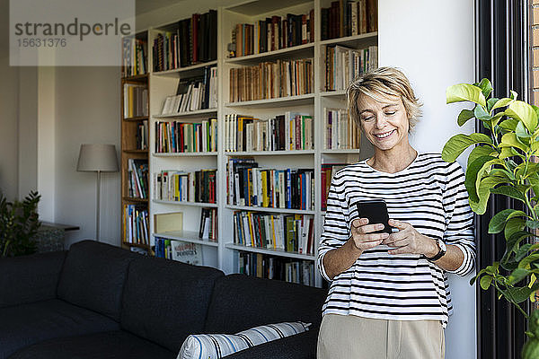 Lächelnde reife Frau  die zu Hause am Fenster im Wohnzimmer steht und ein Smartphone benutzt