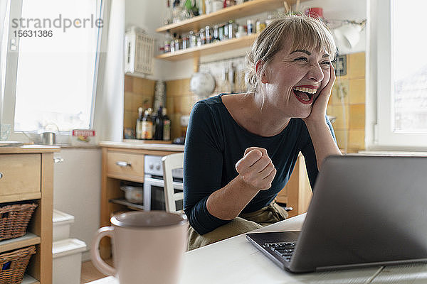 Porträt einer Frau  die mit Laptop in der Küche sitzt und vor Freude weint
