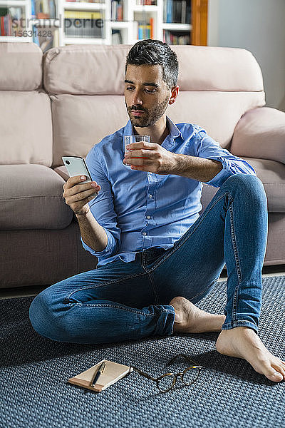 Porträt eines jungen Mannes  der zu Hause barfuss auf dem Boden des Wohnzimmers sitzt und auf ein Smartphone schaut