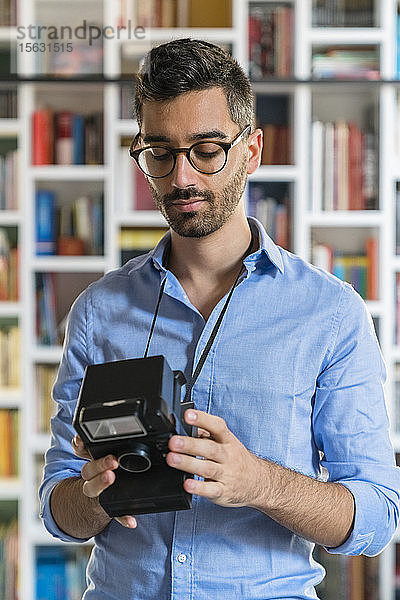 Porträt eines jungen Mannes  der vor einem Bücherregal steht und auf eine Sofortbildkamera schaut