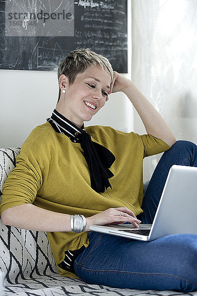 Porträt einer lächelnden Frau  die auf der Couch sitzt und auf den Laptop schaut
