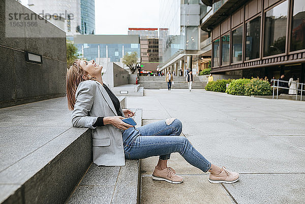Glückliche Frau sitzt mit zurückgelehntem Handy auf einer Treppe in der Stadt