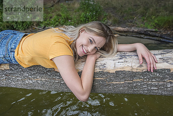 Lächelnde Frau liegt auf einem Baumstamm am Seeufer