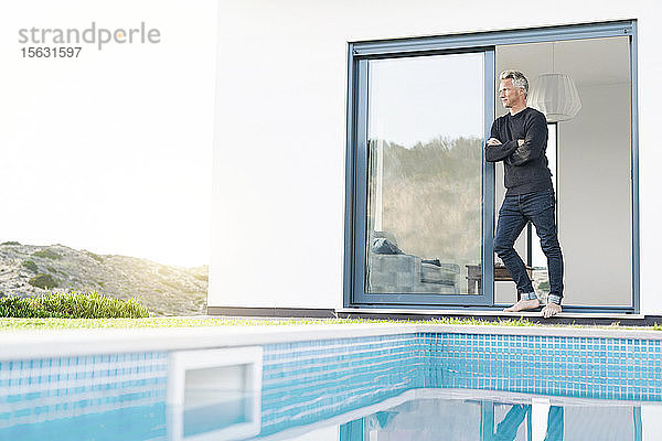 Kluger Mann steht vor seinem modernen Haus mit Pool und schaut in die Landschaft