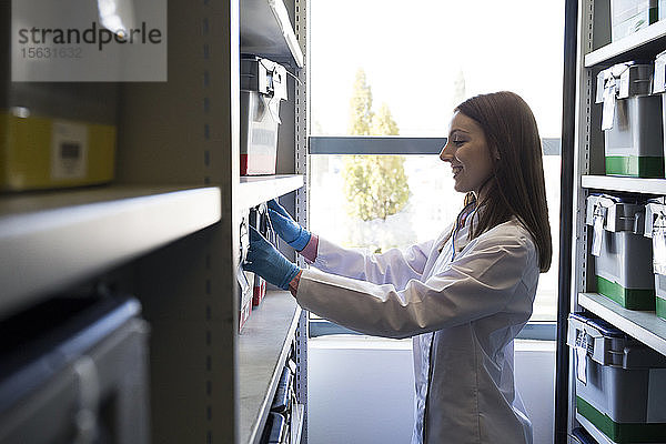 Seitenansicht einer Frau in weißer Robe  die eine Schachtel mit Proben in einem Regal im Labor aufbewahrt