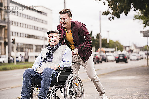 Lachender junger Mann schiebt glücklichen älteren Mann im Rollstuhl