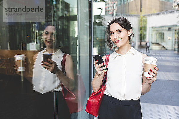 Porträt einer jungen Geschäftsfrau mit Kaffee zum Mitnehmen und Handy  das sich in der Glasfront spiegelt  Berlin  Deutschland
