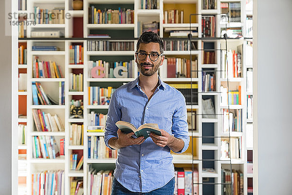 Porträt eines lächelnden jungen Mannes mit Buch  der zu Hause vor den Bücherregalen steht