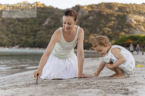 Mutter und Tochter zeichnen mit kleinen Stöckchen im Sand am Strand