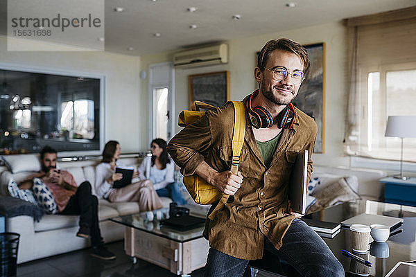Porträt eines lächelnden Mannes mit Rucksack  Kopfhörer und Laptop mit Freunden im Hintergrund