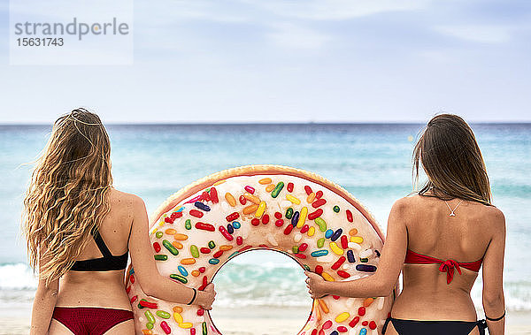 Zwei junge Frauen stehen vor dem Meer und halten einen großen aufblasbaren Ring