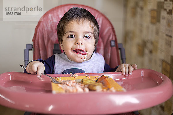 Porträt eines kleinen Jungen  der auf einem Hochstuhl sitzt und Gemüse isst