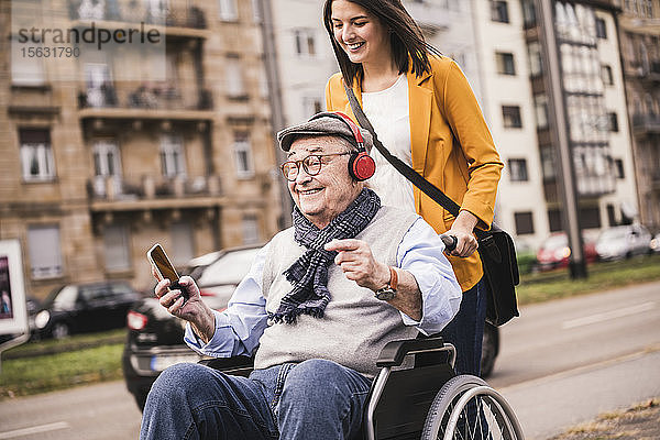 Lachende junge Frau schiebt glücklichen älteren Mann mit Kopfhörern und Smartphone im Rollstuhl