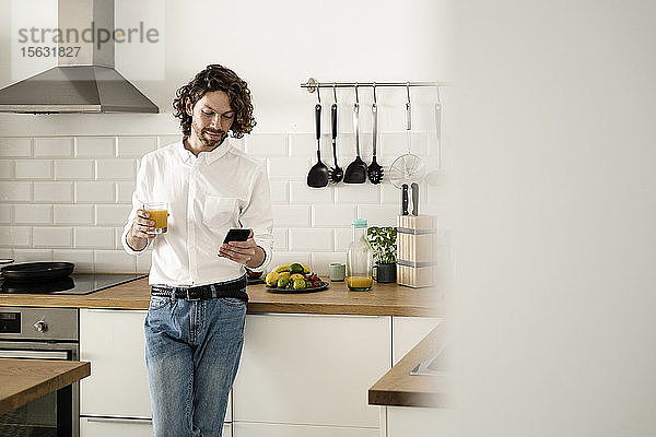 Mann hält zu Hause in der Küche ein Glas Orangensaft und ein Handy