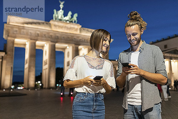 Glückliches Paar benutzt Smartphones am Brandenburger Tor bei Nacht  Berlin  Deutschland