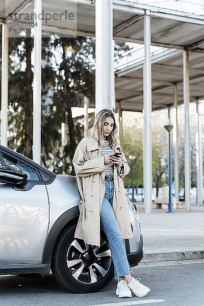 Junge blonde Frau  die ein Smartphone benutzt und sich an ein Auto lehnt