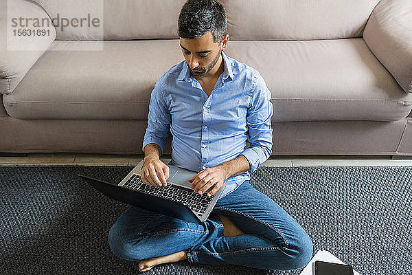 Junger Mann sitzt zu Hause mit Laptop auf dem Boden