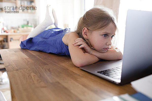 Portrait eines traurigen kleinen Mädchens  das zu Hause auf dem Küchentisch liegt und auf seinen Laptop schaut