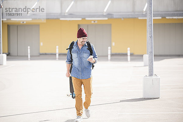 Lässiger reifer Mann mit Smartphone und Skateboard auf dem Parkdeck