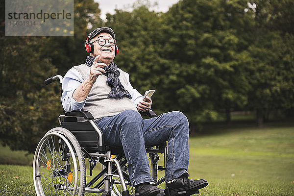Porträt eines glücklichen älteren Mannes  der im Rollstuhl sitzt und mit Smartphone und Kopfhörern Musik hört