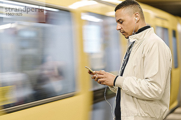 Mann steht mit Kopfhörern und Mobiltelefon am U-Bahn-Bahnsteig  Berlin  Deutschland