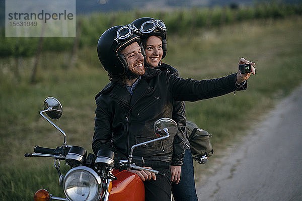 Porträt eines glücklichen jungen Paares auf einem Oldtimer-Motorrad  das sich am Straßenrand ein Selfie gönnt