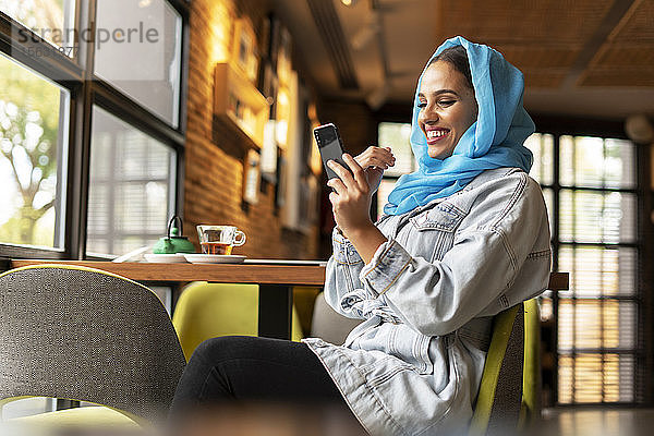 Junge Frau trägt einen türkisfarbenen Hijab und benutzt ein Smartphone in einem Cafe