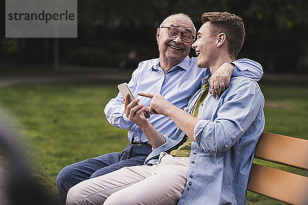 Älterer Mann und Enkel mit Smartphone sitzen zusammen auf einer Parkbank und amüsieren sich