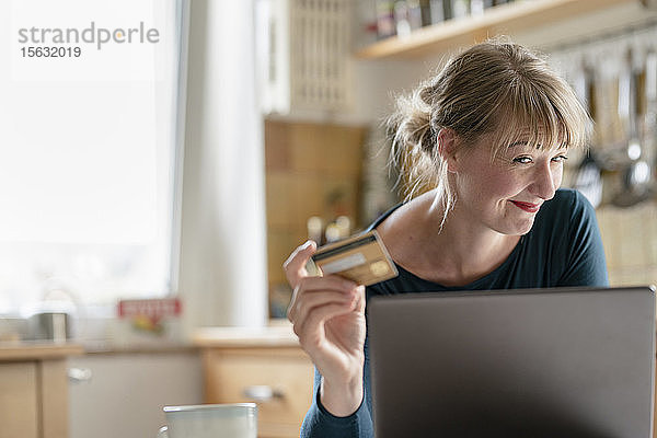 Lächelnde Frau in der Küche mit Kreditkarte und Laptop
