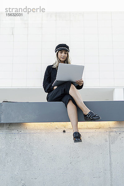Junge blonde Geschäftsfrau mit schwarzer Matrosenmütze und Laptop  die an einer Wand sitzt