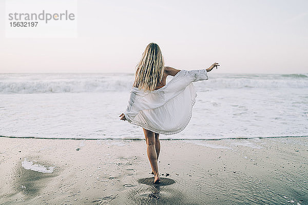 Junge blonde Frau in Bikini und weißem Kleid morgens am Strand