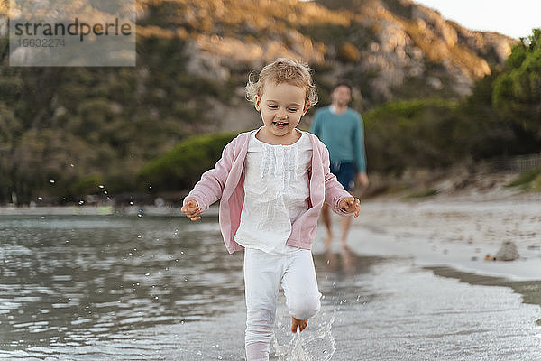 Glückliches Mädchen rennt am Meeresufer mit dem Vater im Hintergrund