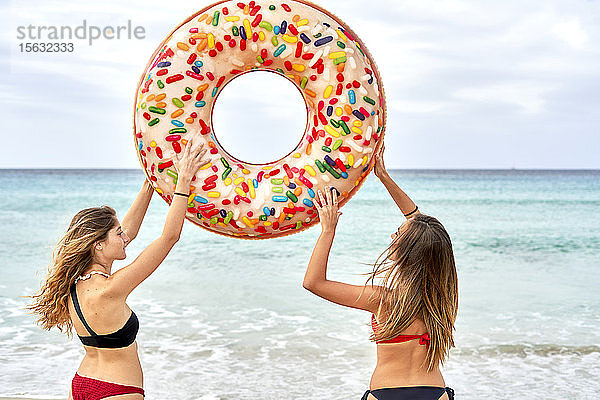 Zwei junge Frauen stehen vor dem Meer und halten einen großen aufblasbaren Ring in der Luft