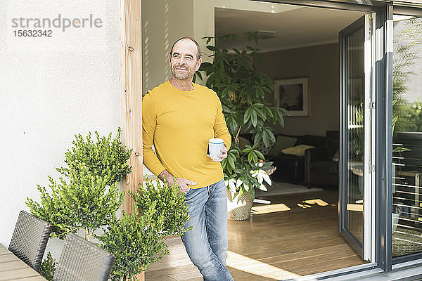 Reifer Mann  der zu Hause mit Kaffeetasse an der Terrassentür steht