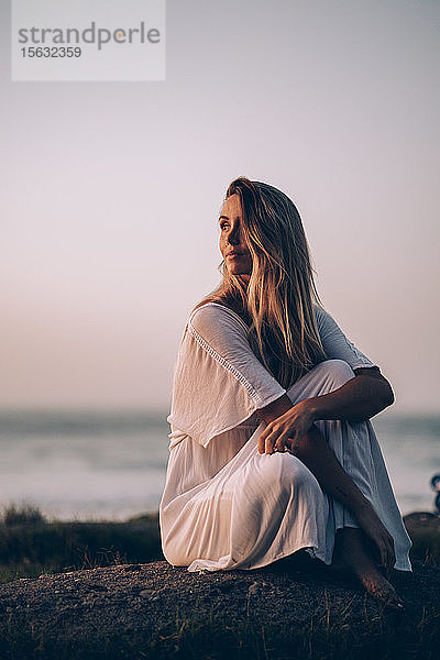 Junge blonde Frau sitzt am Strand und schaut bei Sonnenaufgang zur Seite