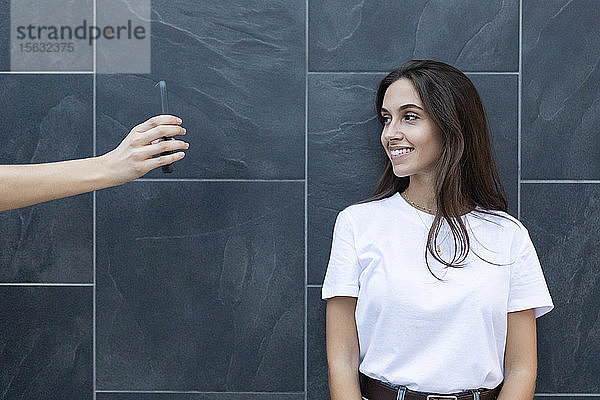 Junge lächelnde Frau schaut auf ein Smartphone  das ihr Freund in der Hand hält  mit einer grauen Wand im Hintergrund