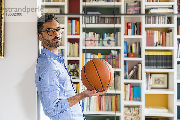Porträt eines jungen Mannes mit Basketball  der zu Hause vor einem Bücherregal steht