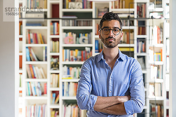 Porträt eines bärtigen jungen Mannes  der zu Hause vor einem Bücherregal steht
