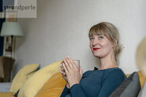 Porträt einer lächelnden Frau  die mit einer Tasse Kaffee auf der Couch sitzt