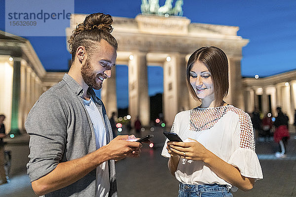 Glückliches Paar benutzt Smartphones am Brandenburger Tor zur blauen Stunde  Berlin  Deutschland